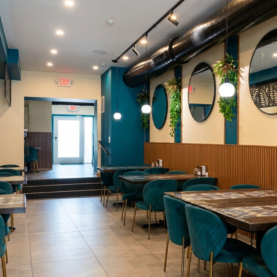 Bright, modern restaurant dining room with teal velvet chairs, marble tables, hanging plants, mirrors, and warm pendant lighting.