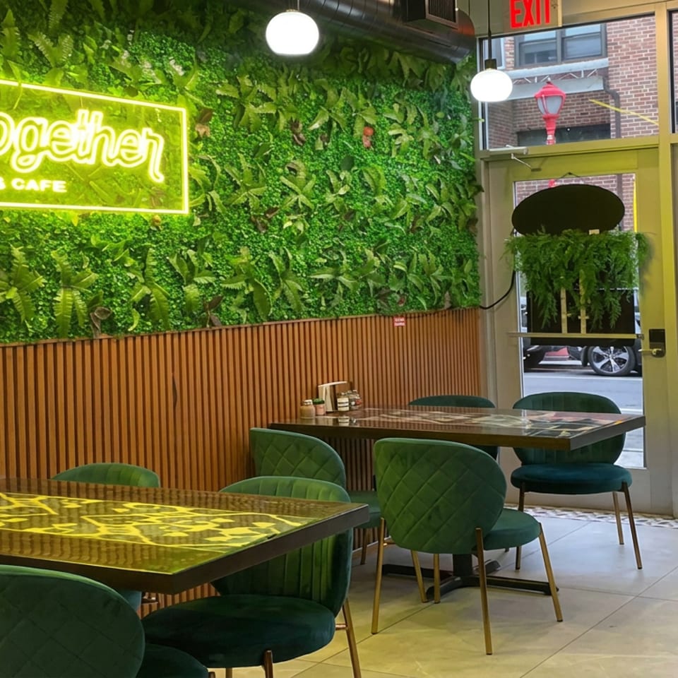 Restaurant café interior with green upholstered chairs, wood-paneled walls, leafy decor, glowing neon sign, and street-facing windows.