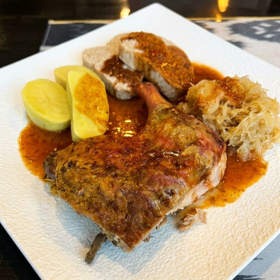 Roasted meat with crispy skin served in sauce alongside potatoes and sauerkraut on a white plate.